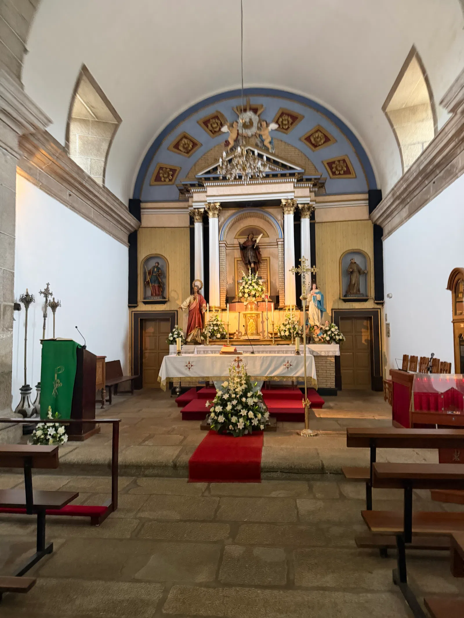 Interior of a church showing altar with statues, floral arrangements, chandeliers, and wooden pews under vaulted ceiling.