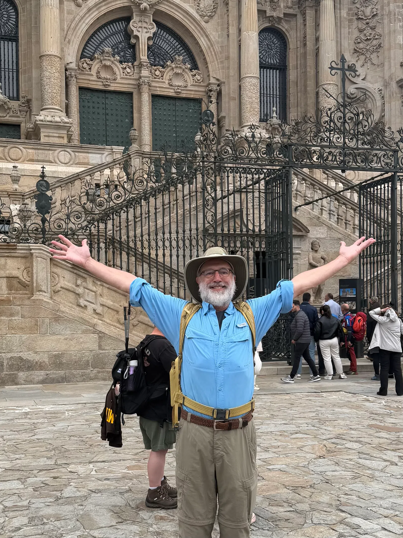 Smiling tourist with open arms in blue shirt and hat in front of ornate metal gate and historic stone building