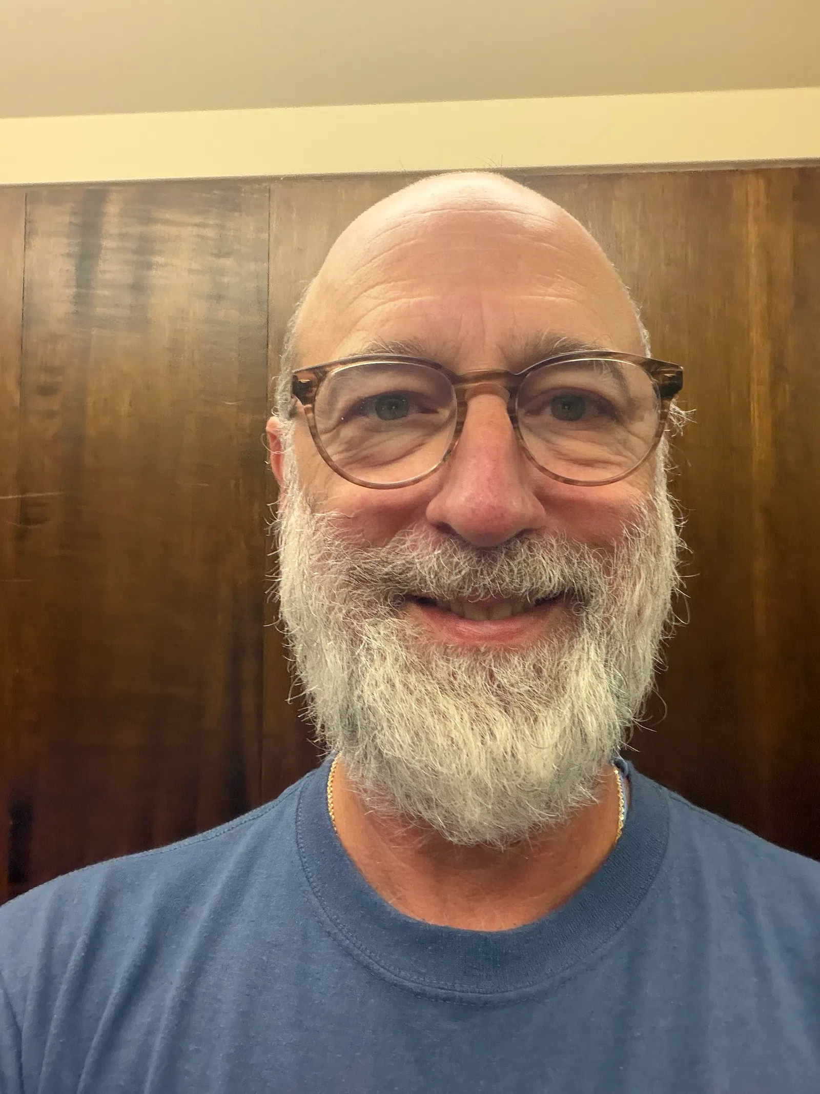 Smiling bald man with white beard and glasses wearing a blue shirt against a wooden background.
