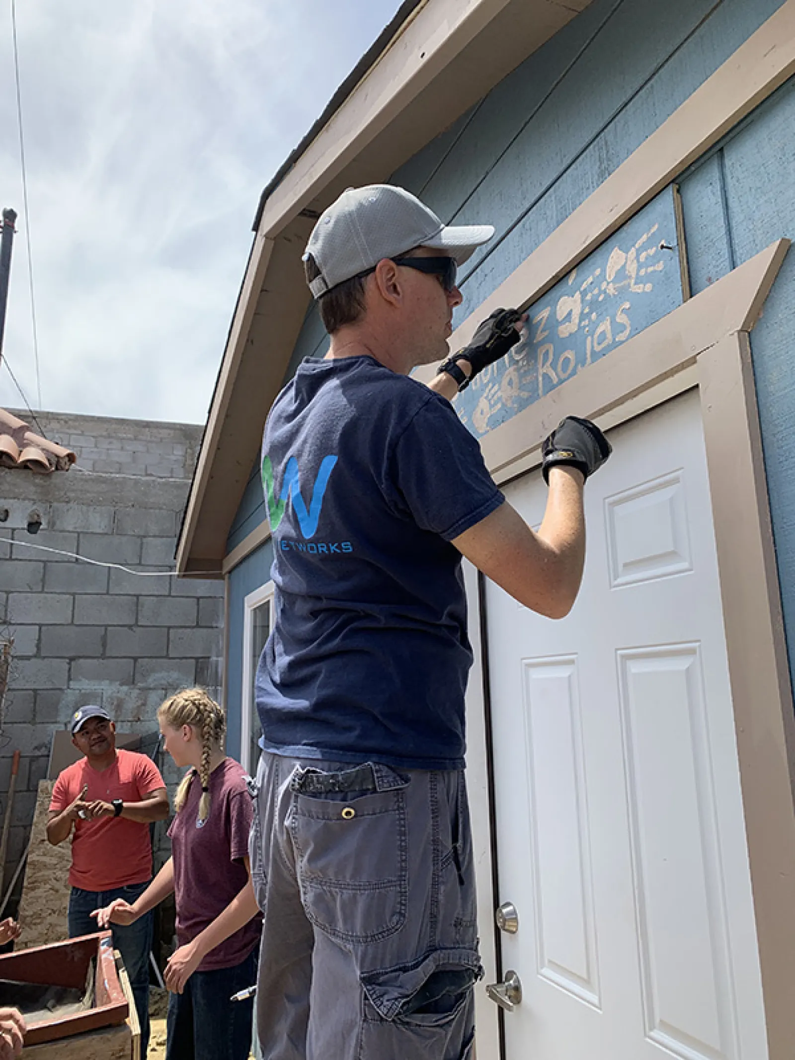 Man wearing cap and gloves painting a wooden sign above a white door on a blue house with two people conversing nearby.