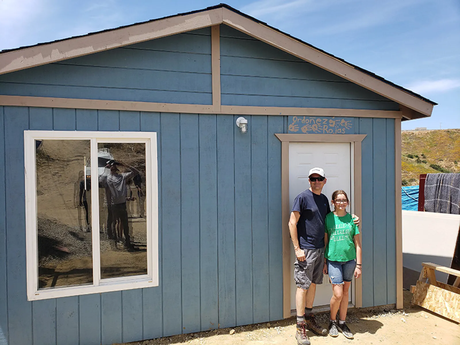 Man and girl standing in front of a small blue wooden house with a white door and window on a sunny day.