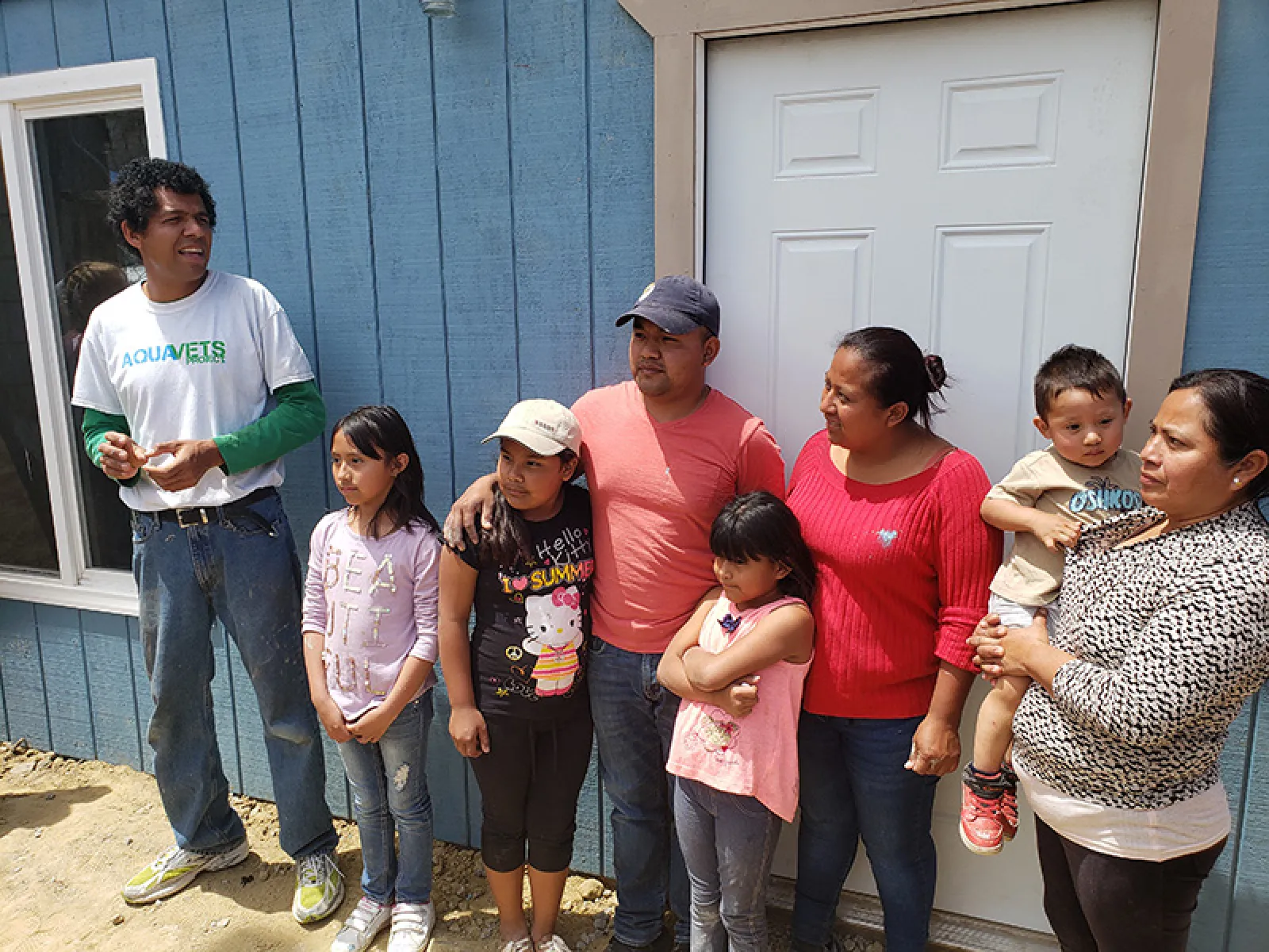 Group of seven people, including adults and children, standing outside a blue house near a white door on a sunny day