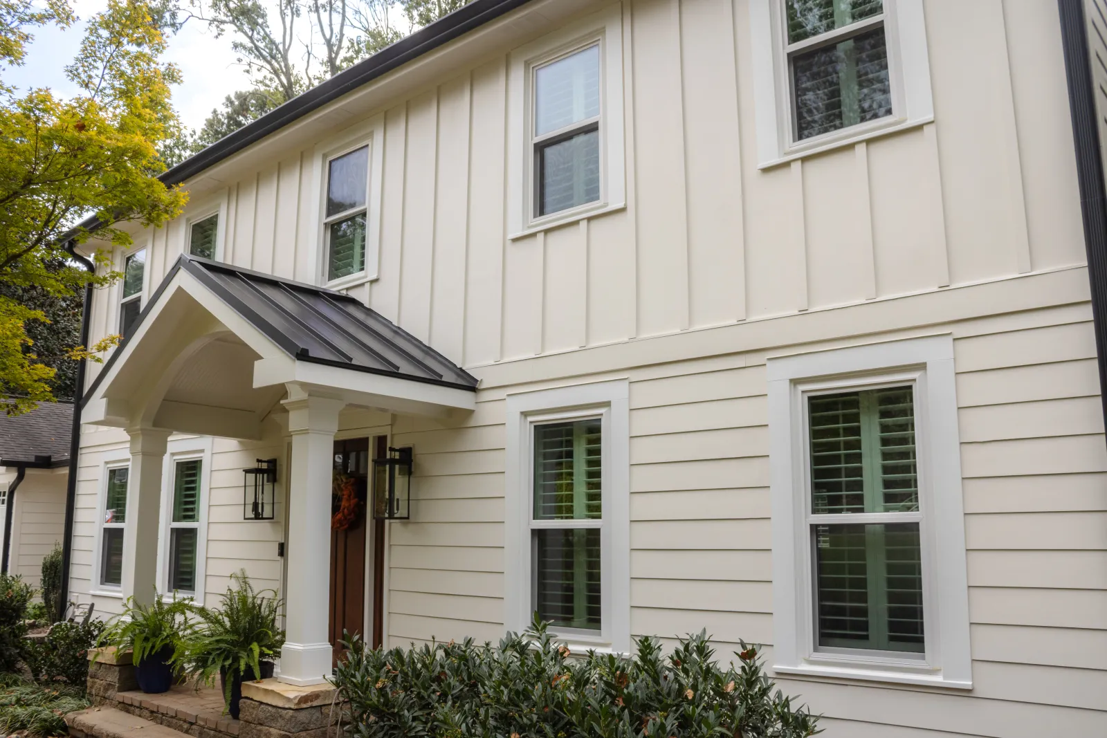 Modern house exterior featuring cream board‑and‑batten siding above horizontal lap siding, white‑framed windows with shutters, dark metal porch roof, and a welcoming front entry with lanterns, columns, and potted plants surrounded by greenery in Atlanta
