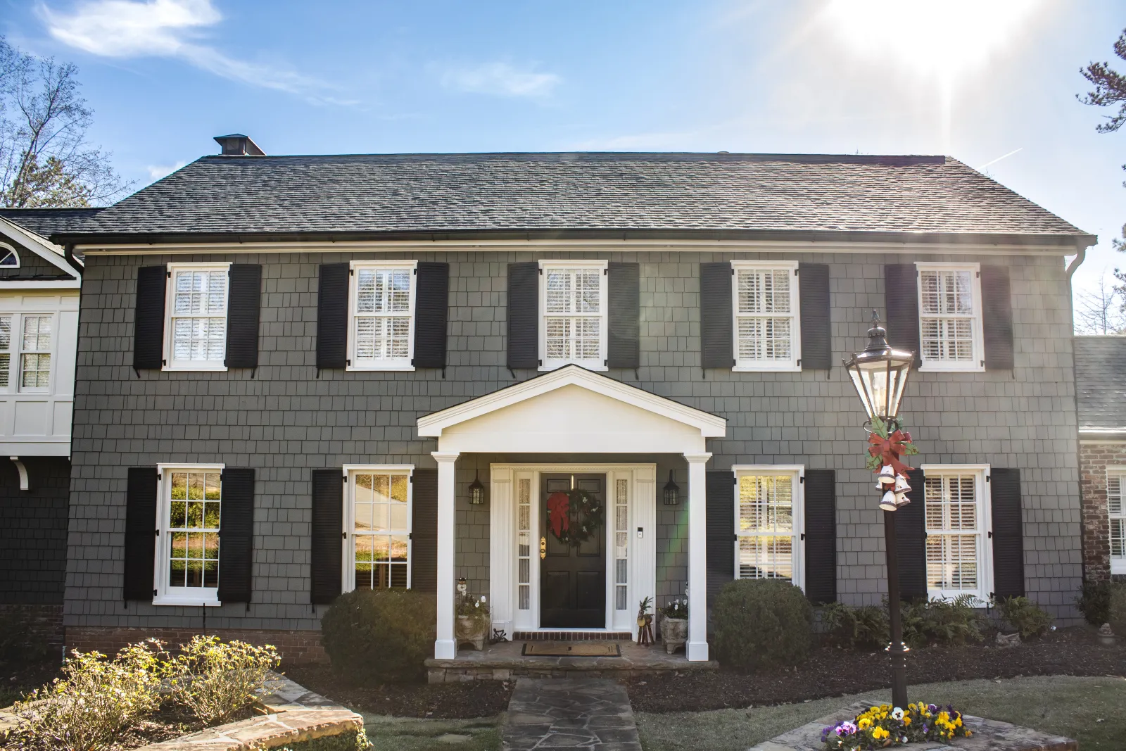 Front exterior of a two‑story house with dark gray shingle siding, white trim, and black shutters, featuring a portico entrance with wreath‑decorated door, landscaped walkway, and a festive lamp post with red bow and bells  in Marietta