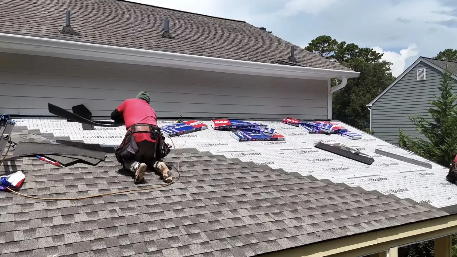 Nelson Exterior Employee kneeling on a roof installing asphalt shingles over underlayment labeled FeltBuster, with GAF Timberline HD shingle bundles, tools, and nails visible nearby in Marietta