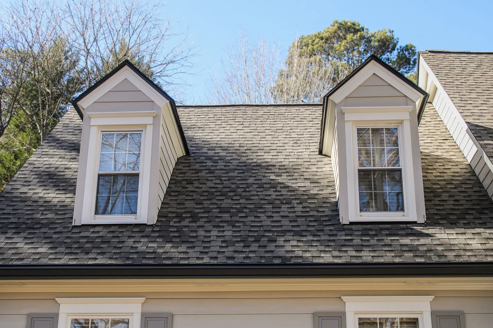Roof with two white dormer windows on a house with gray shingles and clear blue sky background.