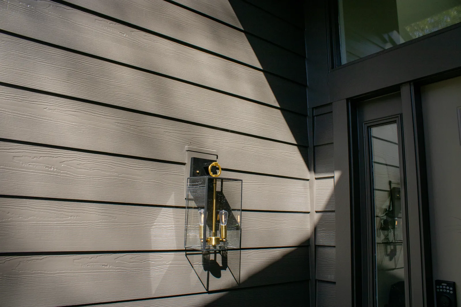 Modern exterior wall with horizontal gray siding, featuring a brass and glass outdoor light fixture casting a shadow, next to a door with vertical glass panel and keypad lock in Atlanta