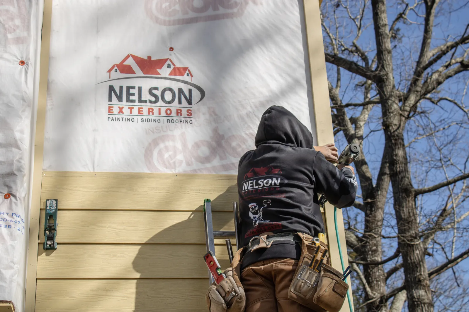 Nelson Exteriors employee installing beige horizontal siding with a nail gun, wearing branded hoodie and tool belt, beside a ladder and house wrap displaying company logo in Atlanta