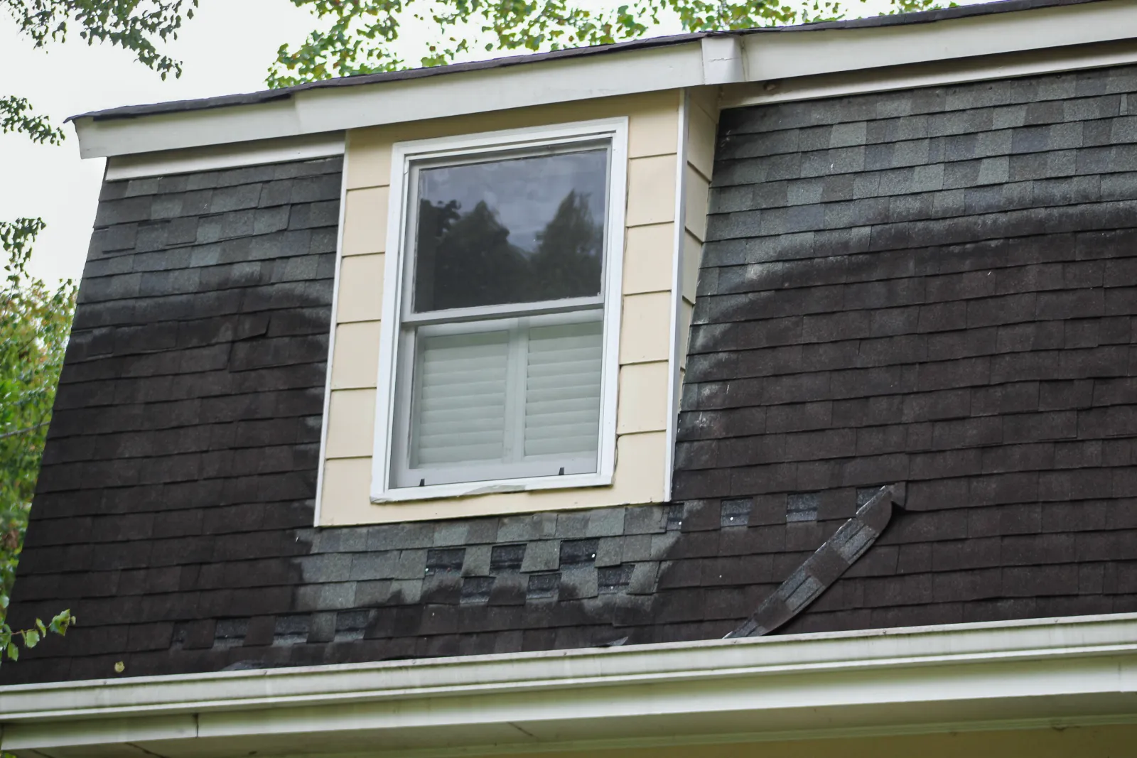House roof with asphalt shingles showing discoloration from algae or moisture buildup and missing or damaged shingles near the lower edge, with a dormer window framed in white trim in Marietta