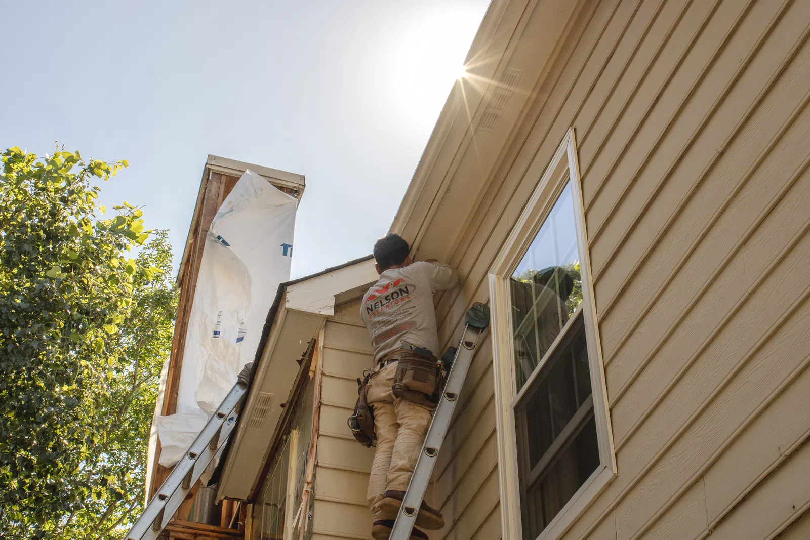 A contractor wearing a Nelson Exteriors shirt standing on an extension ladder to work under the roof soffit of a beige two-story house, installed by Marietta deck builders, home repair in Marietta by Nelson Exteriors.