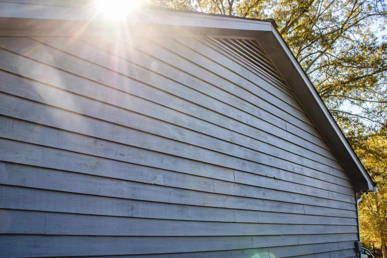 Side view of a building with light gray wooden siding and gable roof, sunlight reflecting off the panels with lens flare, framed by trees with green and yellow leaves in Marietta