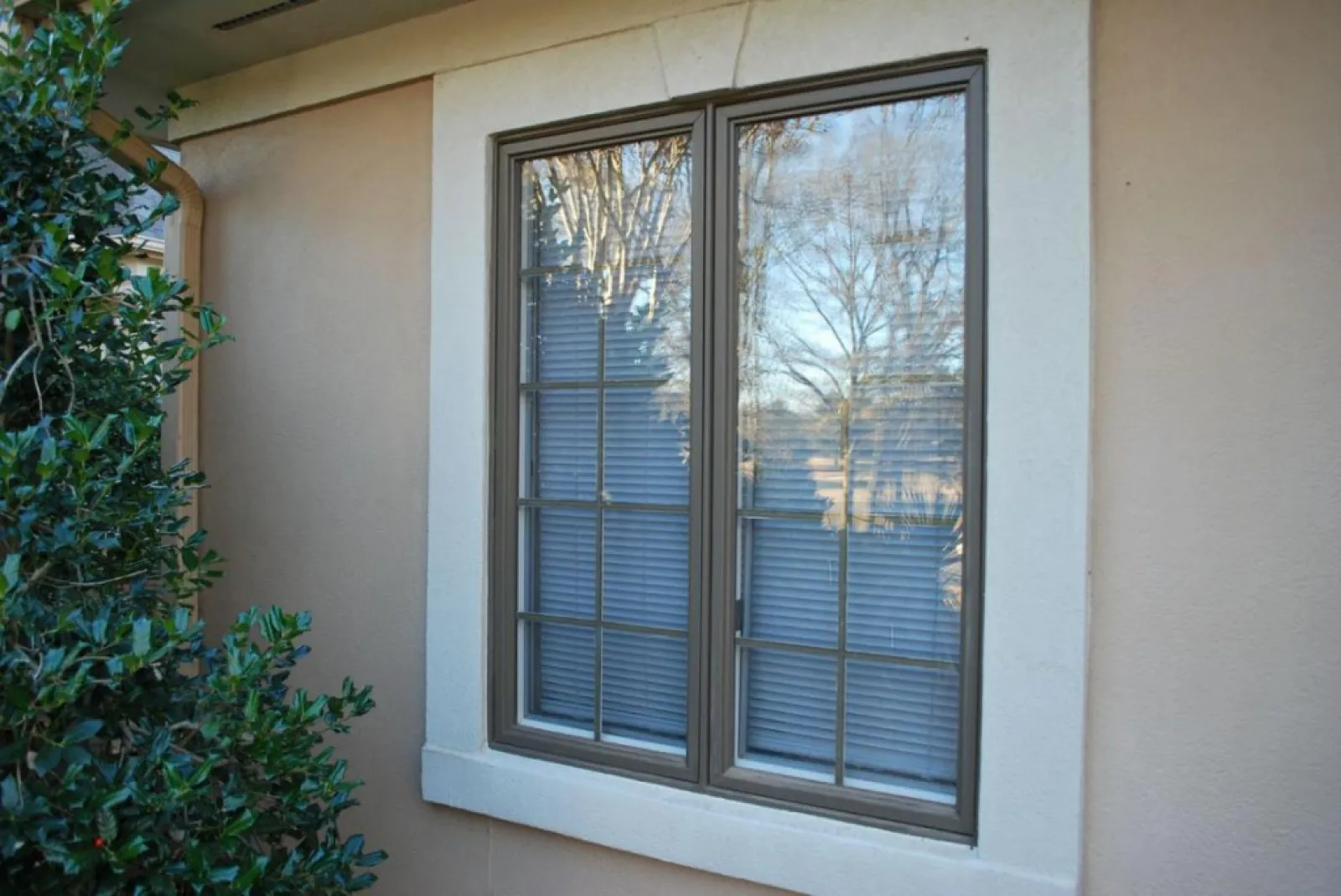 Reflective double-pane window with blinds on beige stucco wall next to green shrubbery outside a residential home.