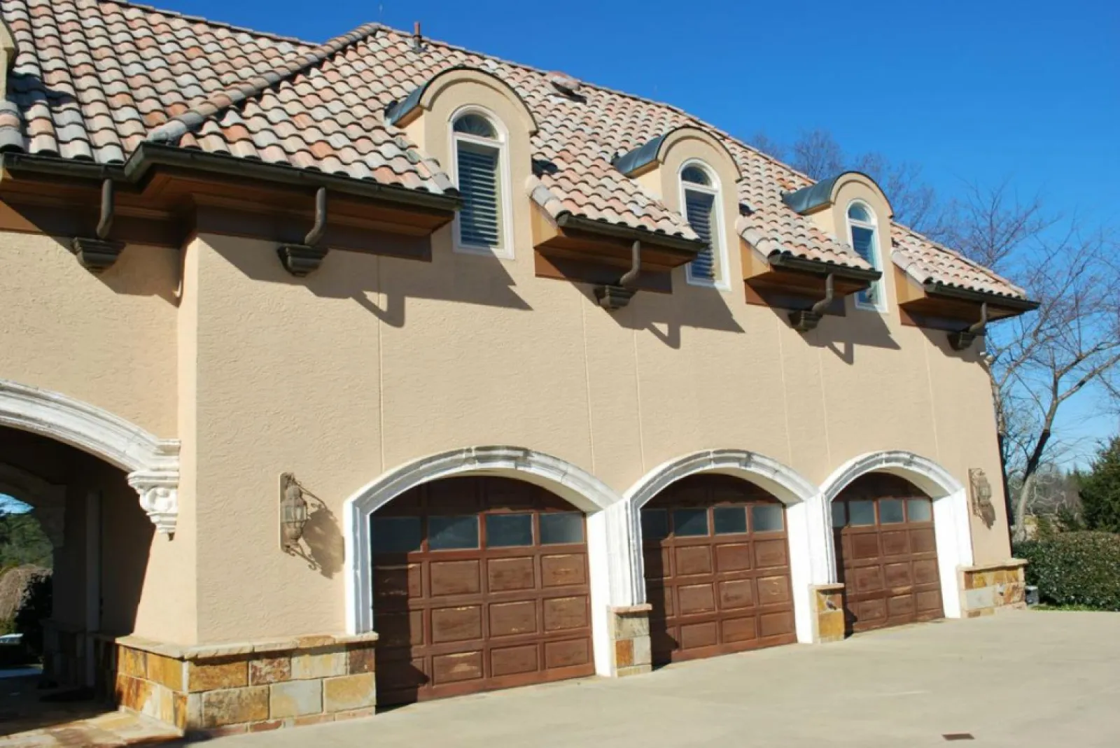 Mediterranean-style house with three wooden garage doors and arched windows under a tiled roof.