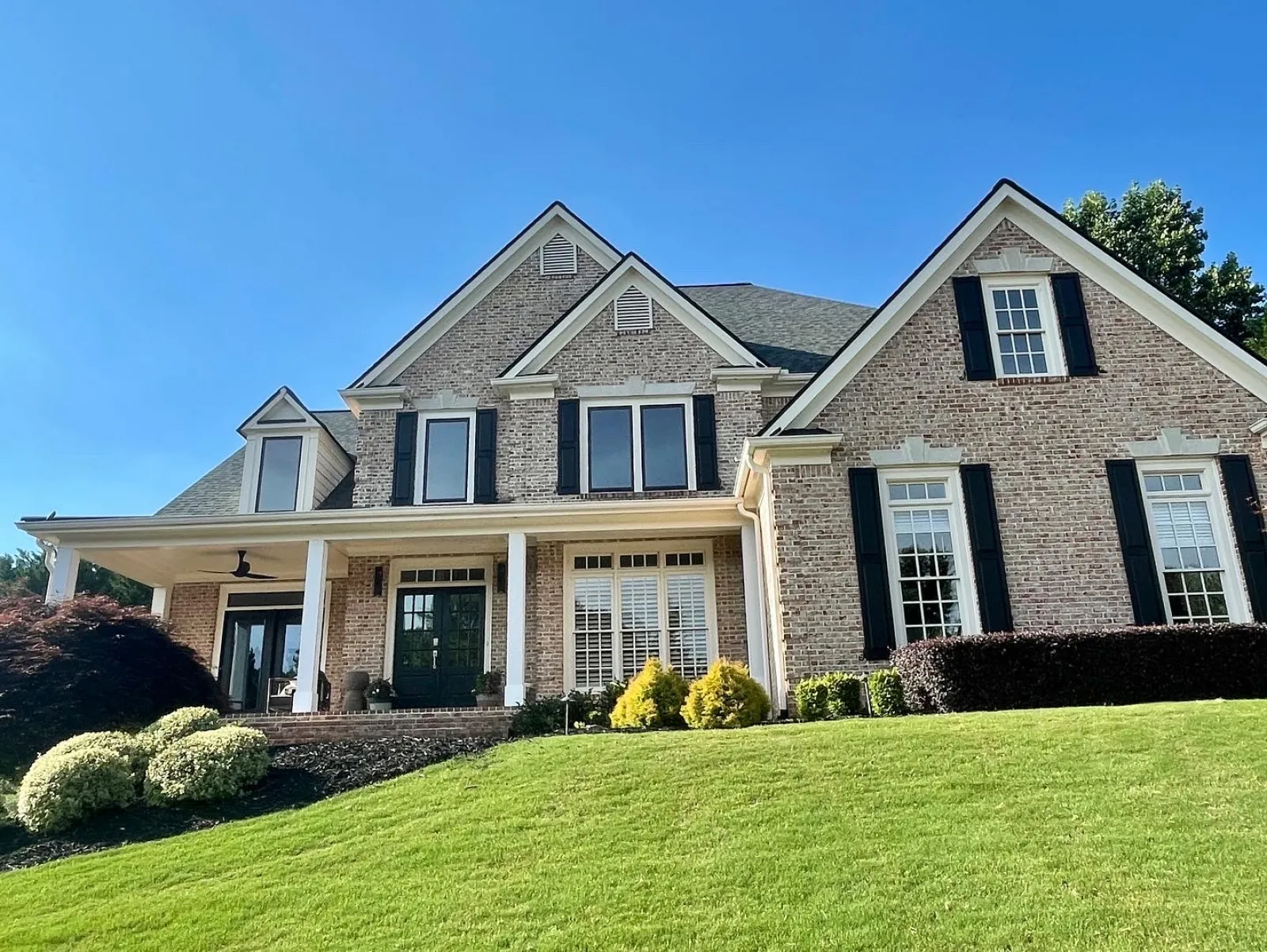 Large brick suburban house with black shutters and a manicured green lawn under a clear blue sky.