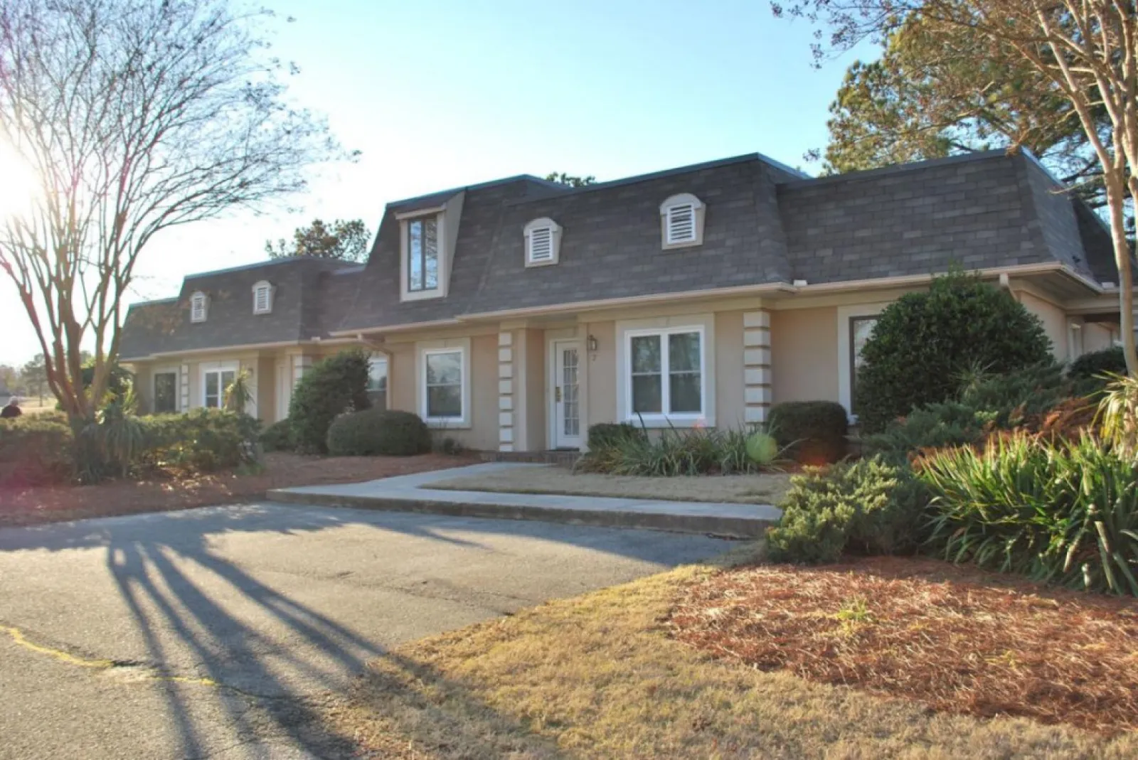 Single-story beige house with dormer windows, surrounded by bushes and trees in a sunny suburban setting.