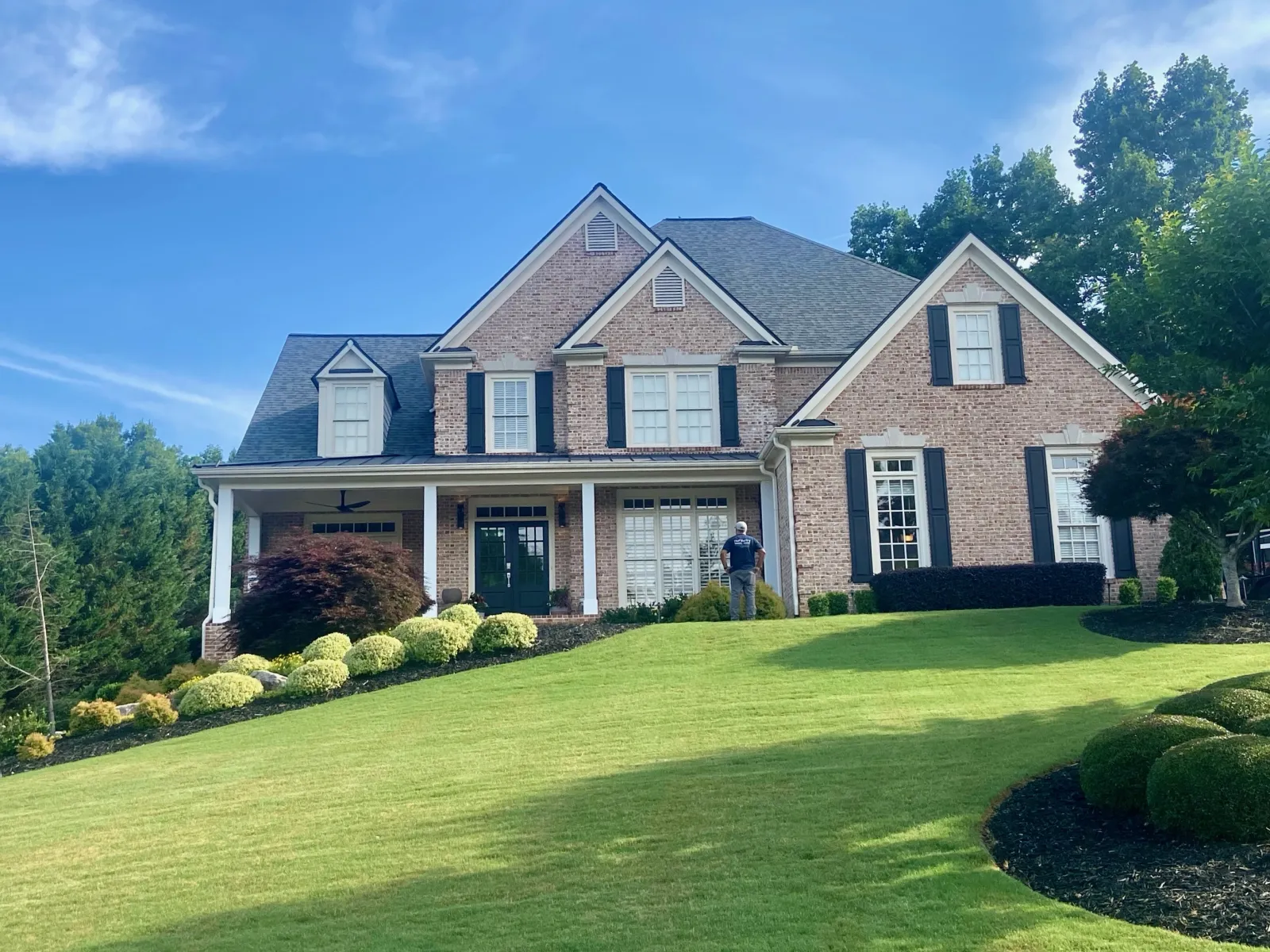 Large brick two-story house with black shutters, manicured green lawn, and trees under a blue sky.