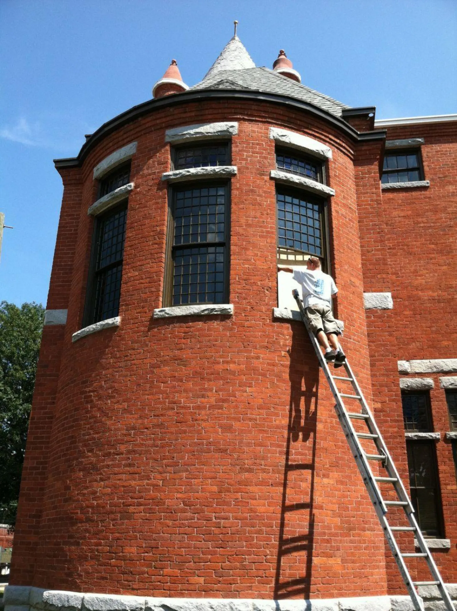 Man on ladder painting or cleaning a large curved red brick building with tall windows under a clear blue sky