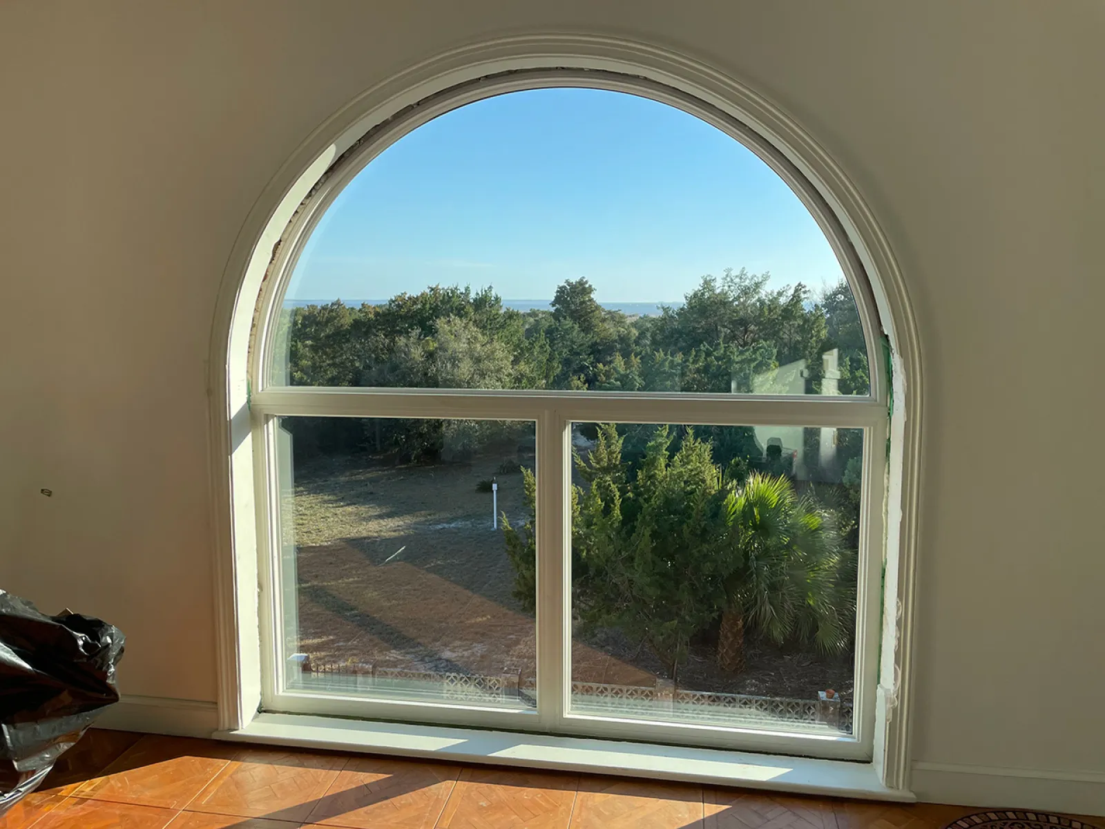Arched window with white frame overlooking trees and blue sky with sunlight casting shadows on hardwood floor.