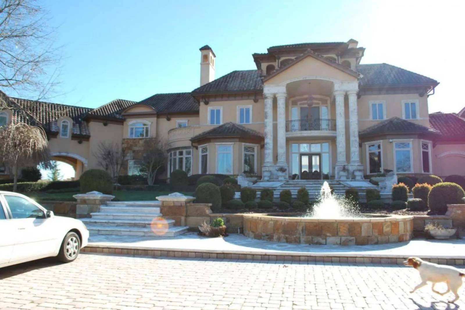 Spacious luxury mansion with classical columns, landscaped garden, stone fountain, and a white car parked outside.