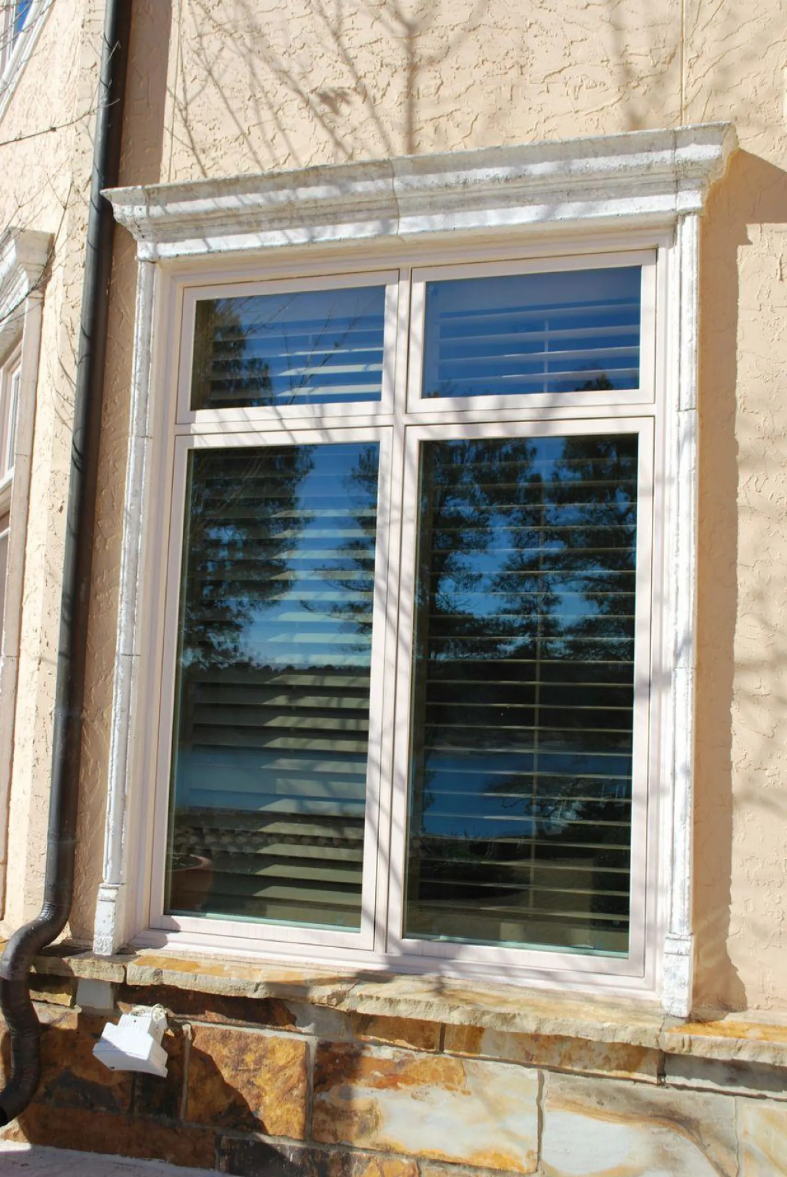 Modern window with white shutters and stone window sill on beige stucco wall reflecting trees and sky.