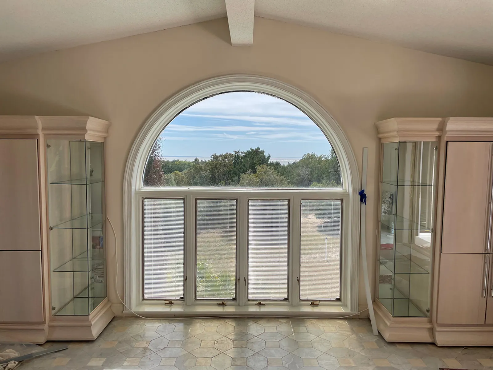 Bright room with large arched window, beige cabinets, glowing tiled floor, and outdoor greenery view.
