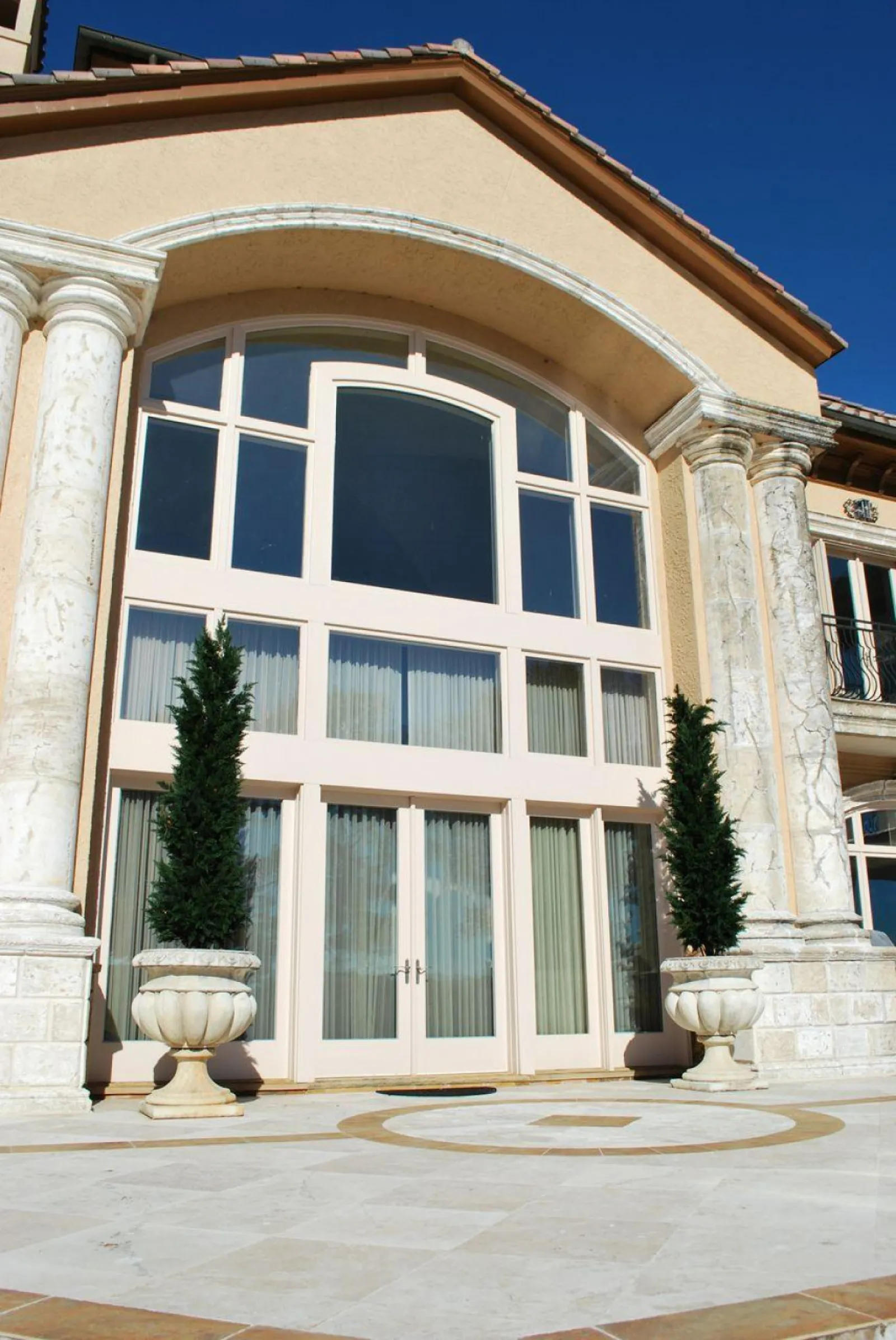 Grand entrance with tall arched windows, marble columns, and potted evergreen trees under clear blue sky.