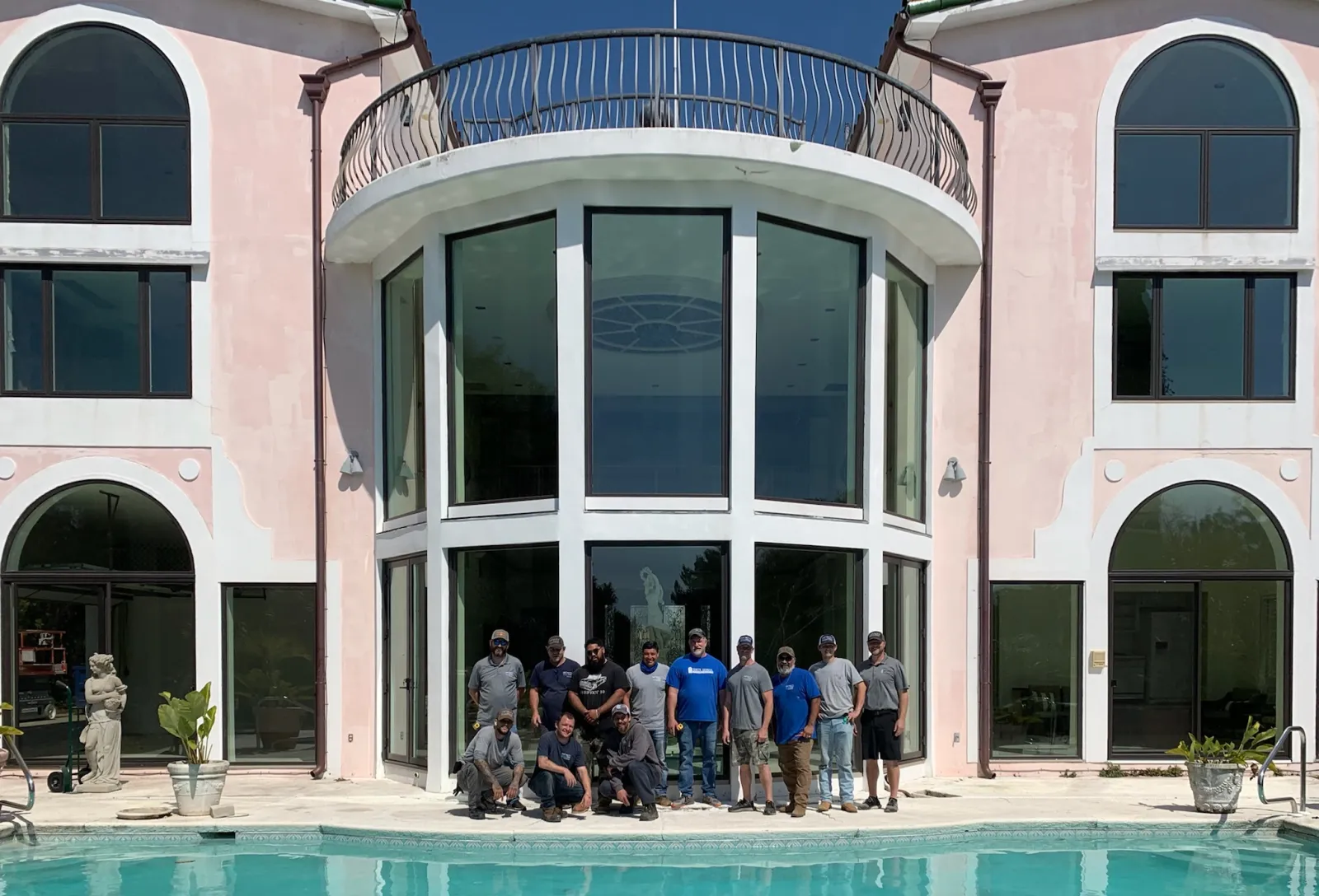 Group of people posing by a large modern house with tall windows and a swimming pool in front.