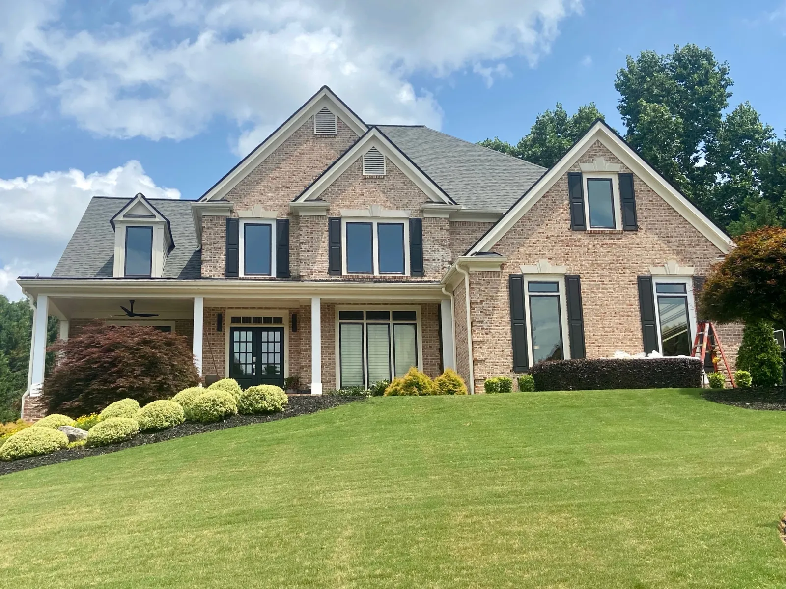 Spacious two-story brick house with black shutters and well-maintained front yard under a blue sky with clouds