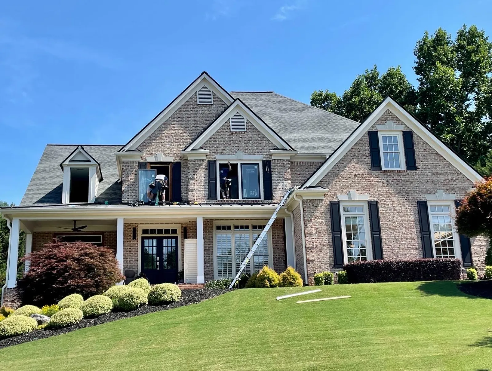 Brick two-story house with black shutters undergoing window installation on a sunny day with green lawn.