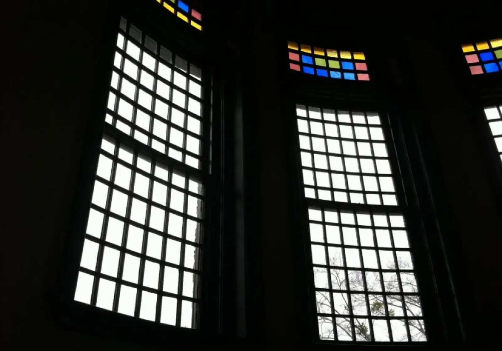 Tall windows with grid patterns and colorful stained glass panels at the top in a dark interior setting