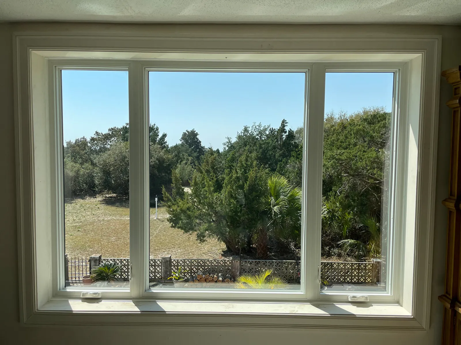 View through a large three-panel window showing trees, bushes, and a clear blue sky outside a fenced yard.