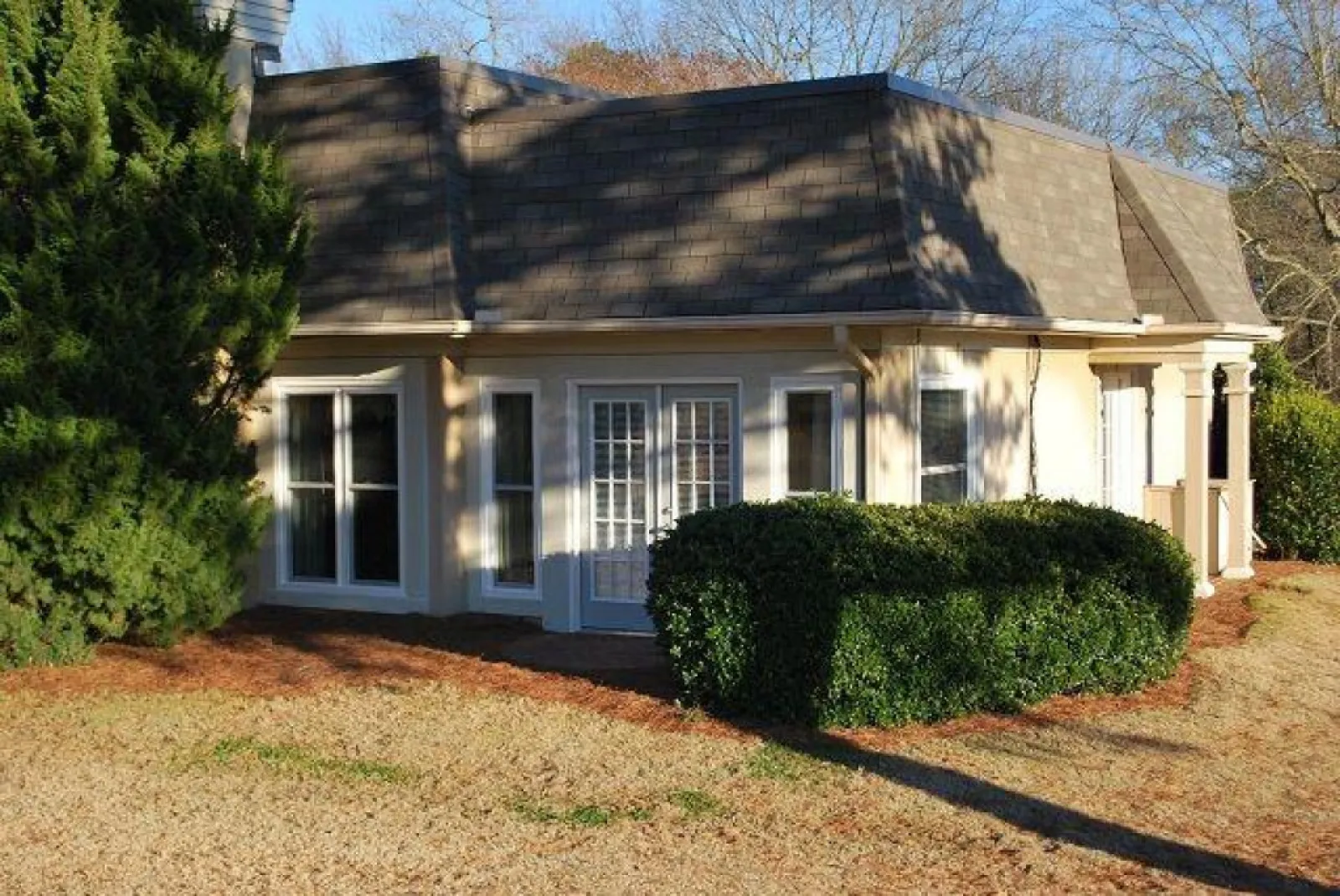 Small beige house with dark roof surrounded by trees and bushes on a sunny day.
