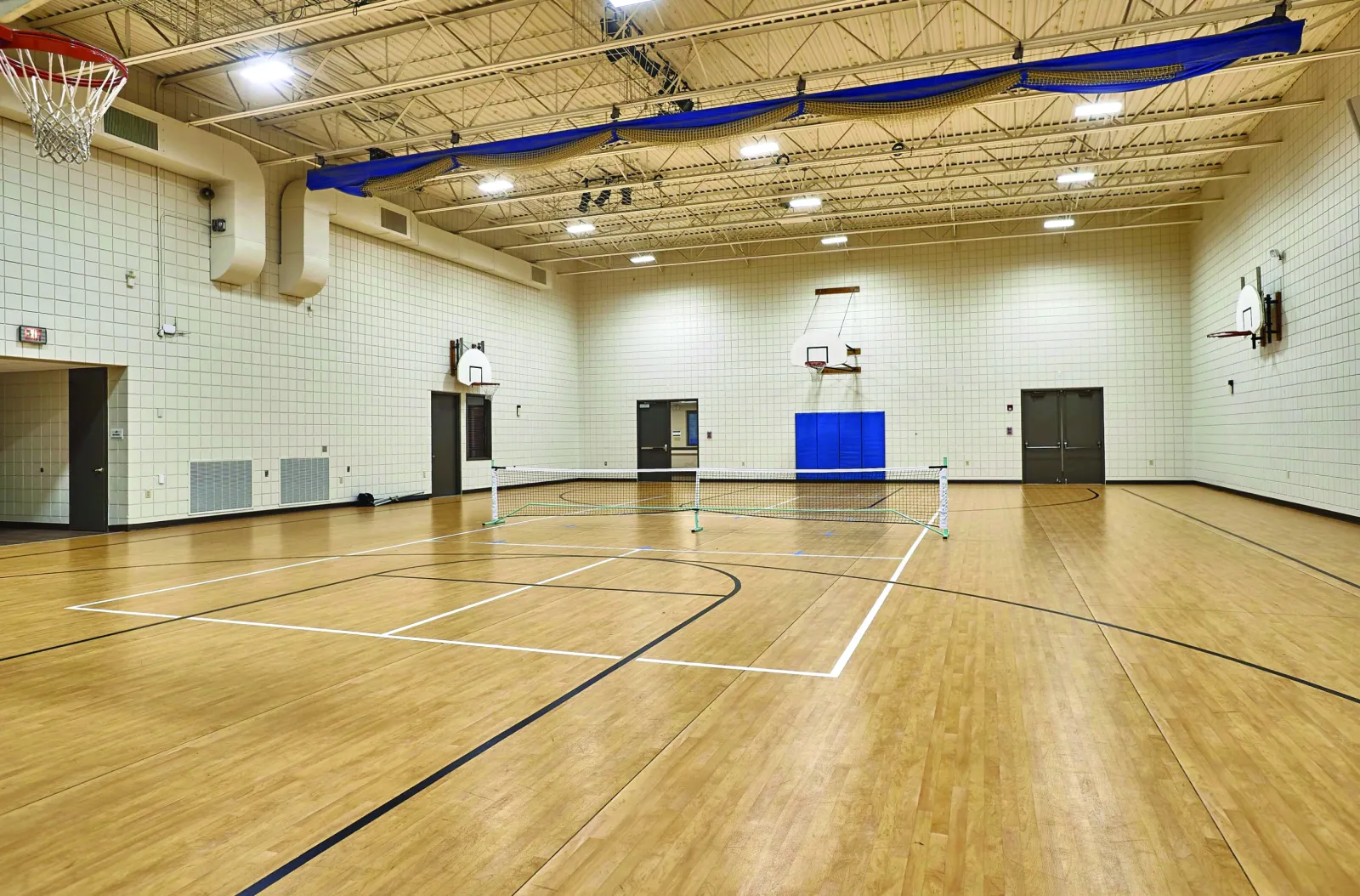 An empty indoor gym with a tennis court, basketball hoops, and wooden flooring.