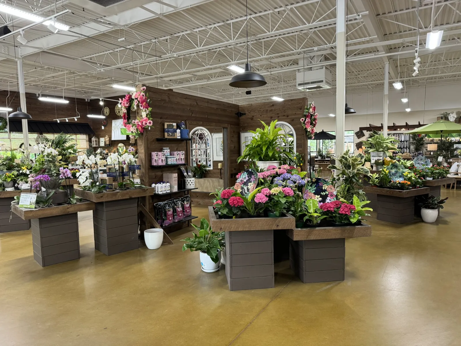 Bright indoor garden center with tables displaying colorful flowers, plants, and garden supplies in a spacious room.