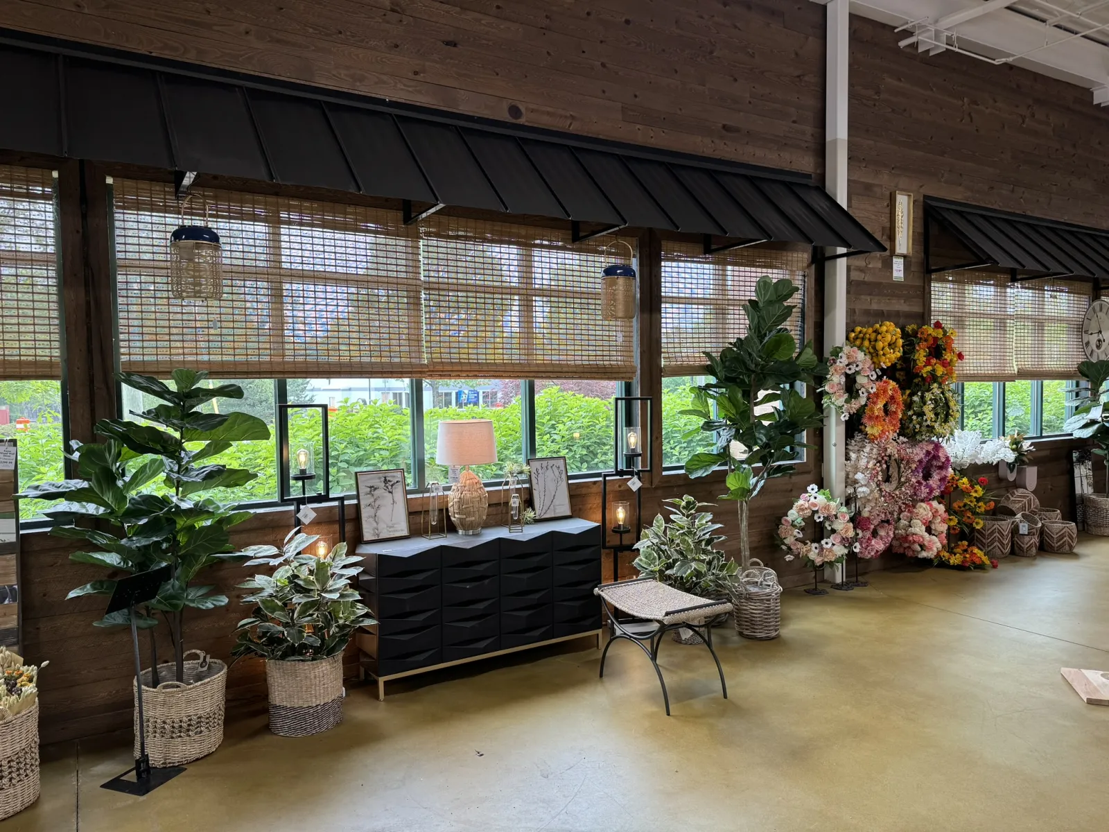Indoor garden shop display with potted plants, woven baskets, floral arrangements, and a black storage cabinet under wooden windows.