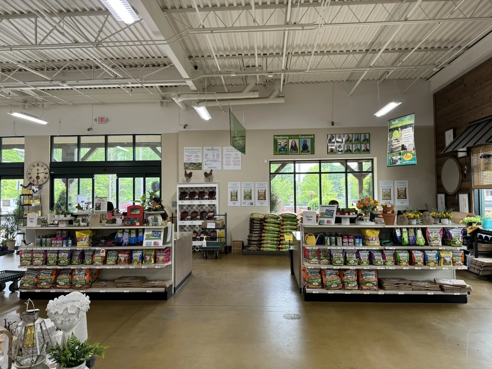 Interior of a bright gardening store with shelves of seeds, soil bags, gardening tools, and plants near large windows.