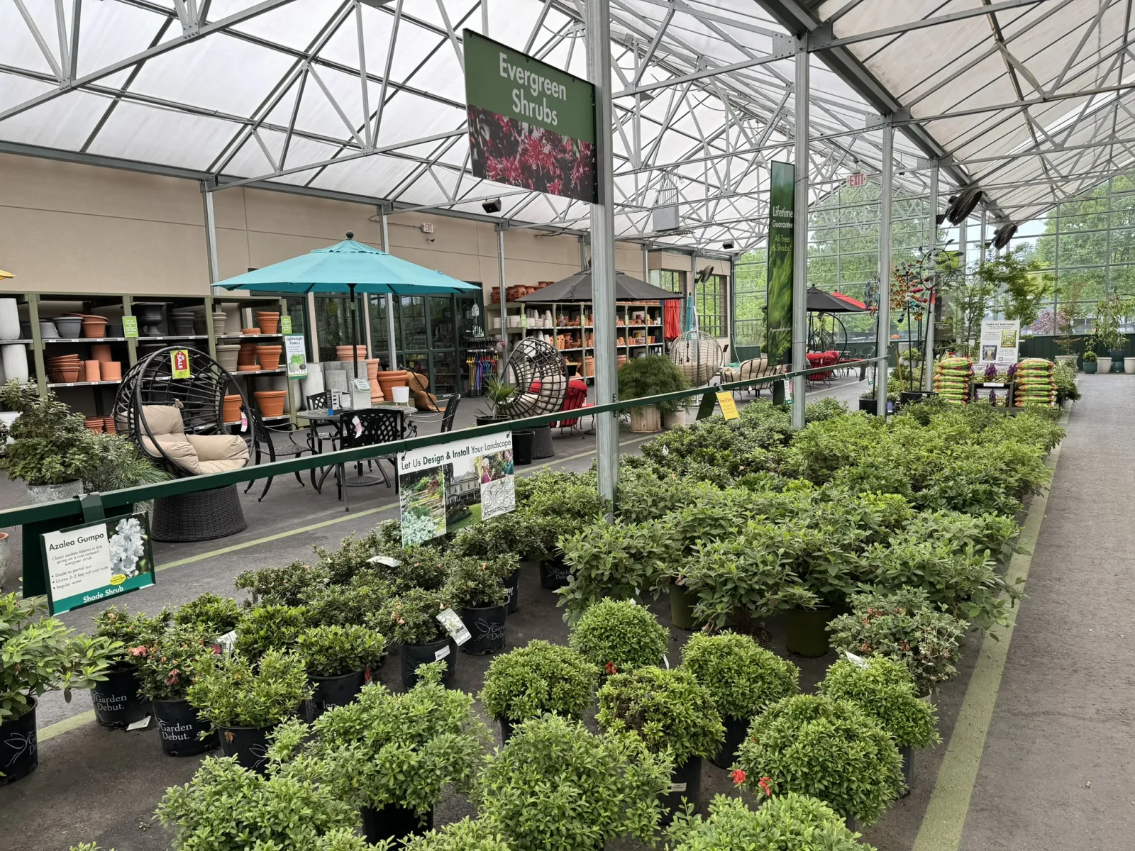 Indoor garden center with rows of evergreen shrubs, potted plants, patio furniture, and gardening supplies under a large greenhouse roof.