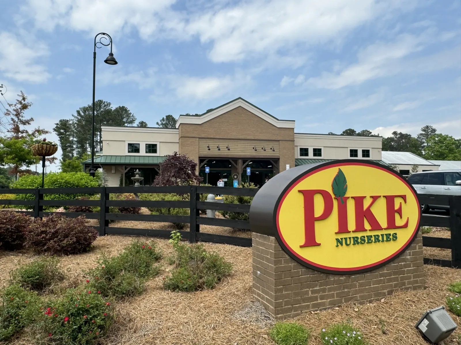 Pike Nurseries sign in front of a landscaped garden and a brick building under a partly cloudy sky.