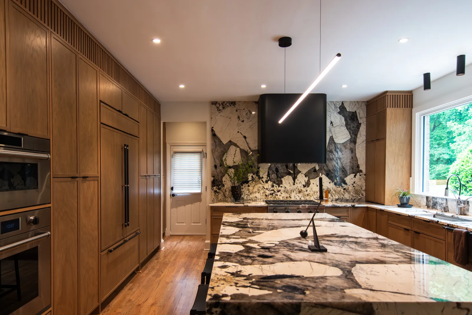 Modern kitchen with large patterned marble island, wooden cabinets, stainless steel appliances, and large window.