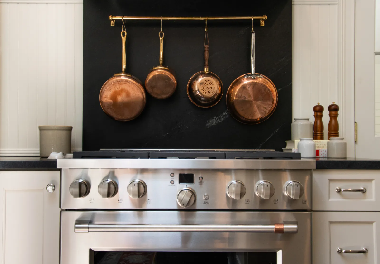Stainless steel stove with control knobs and four hanging copper pots against black backsplash in modern kitchen.