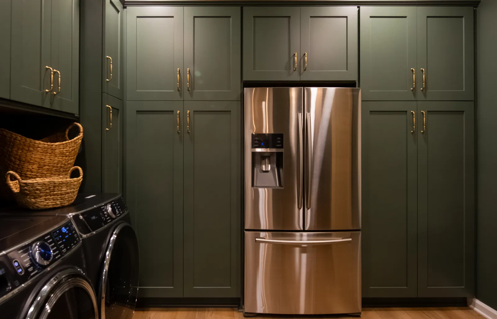 Modern laundry room with olive green cabinets, stainless steel refrigerator, washer, dryer, and woven baskets.