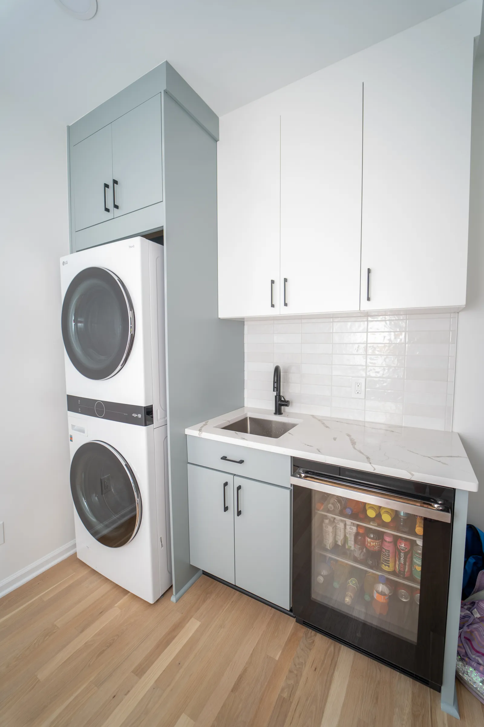 Modern laundry room with stacked washer and dryer, gray cabinets, white countertop, sink, and beverage fridge.
