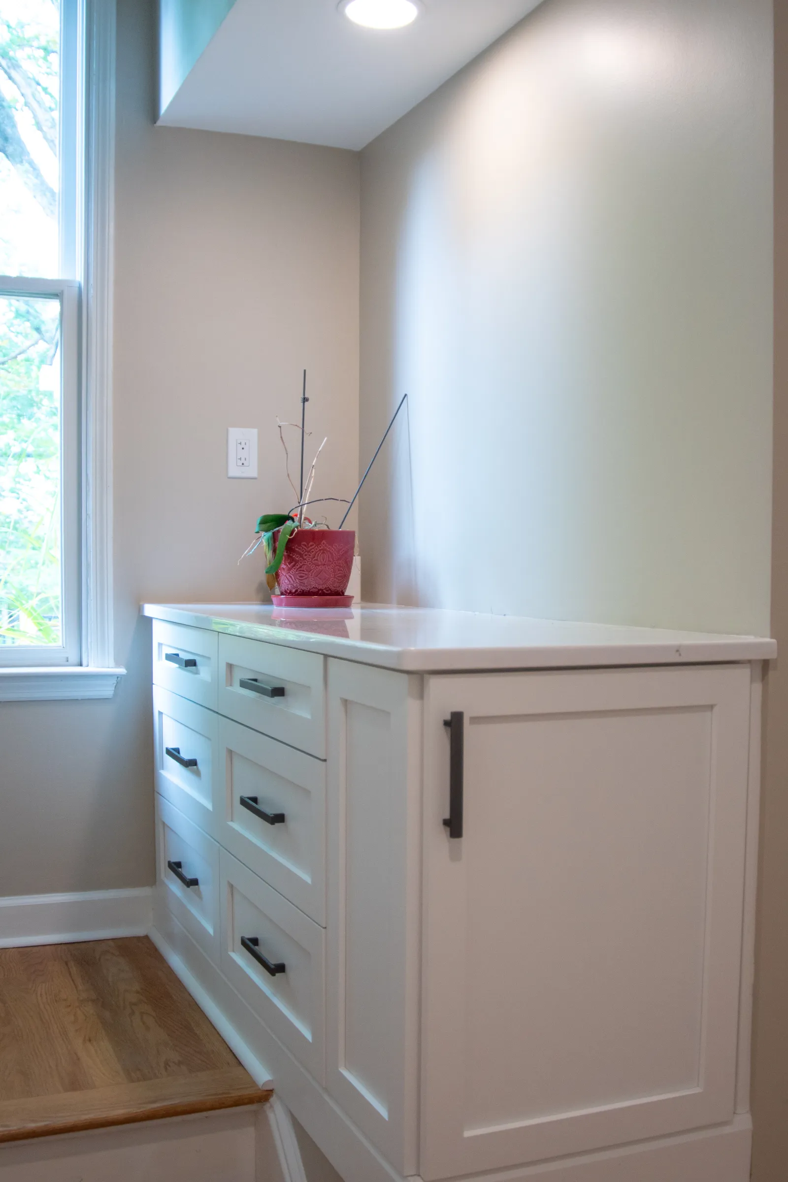 White kitchen cabinetry with black handles beside a window and a small plant on the countertop under recessed lighting.