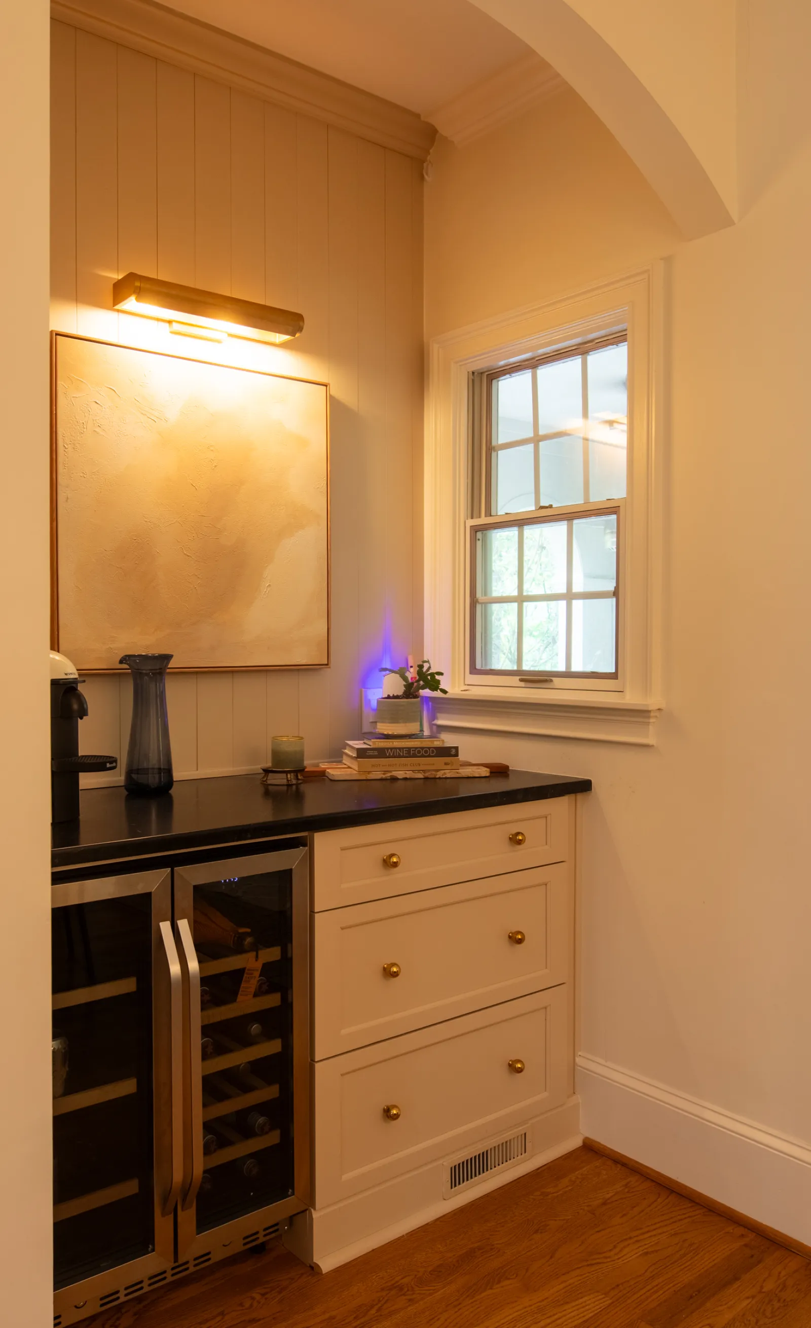 Cozy kitchen corner with black countertop, wine fridge, cream cabinets, decorative art, and window letting natural light in.