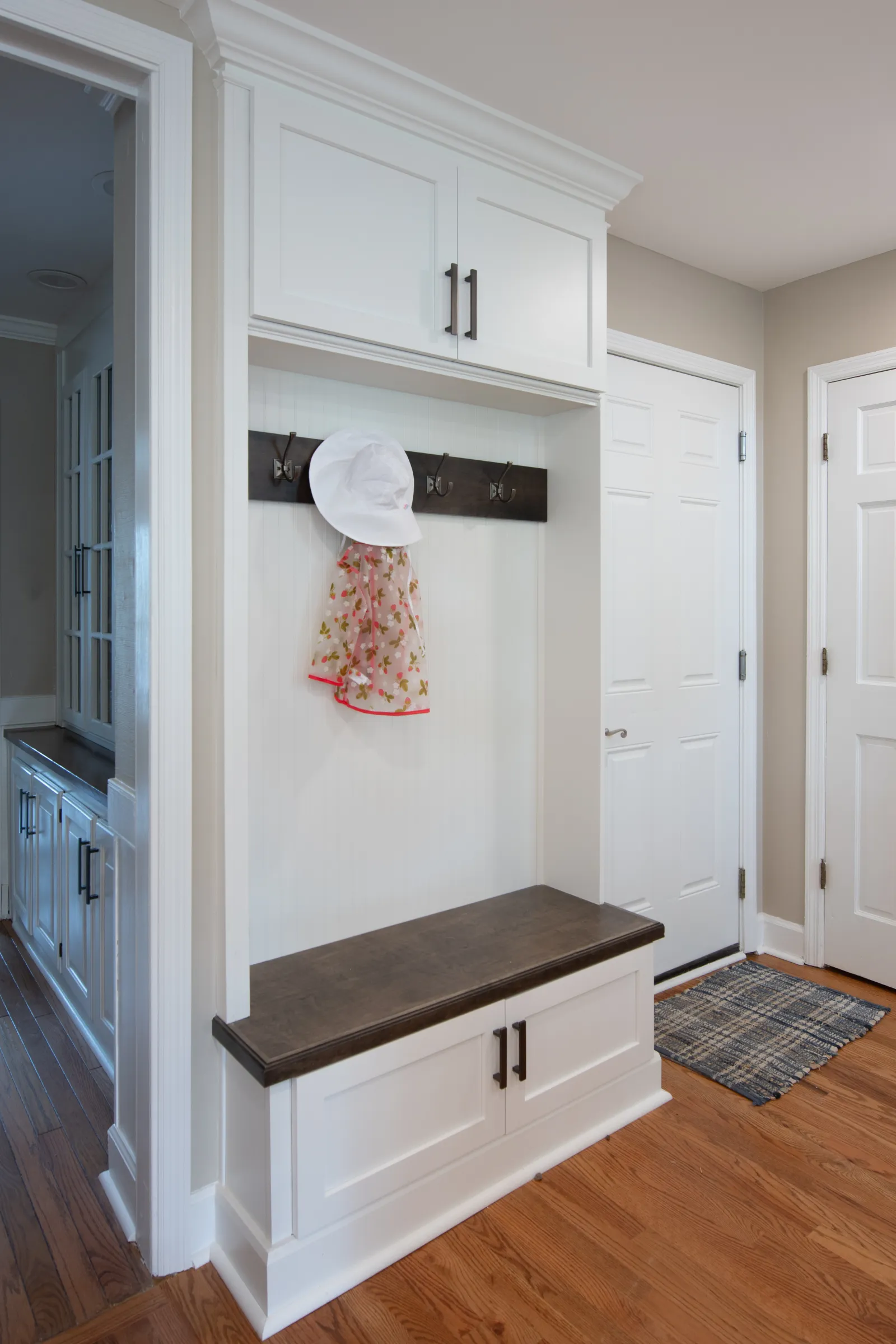 White entryway bench with dark wood top, coat hooks holding a hat and scarf, next to white doors and wood floors.