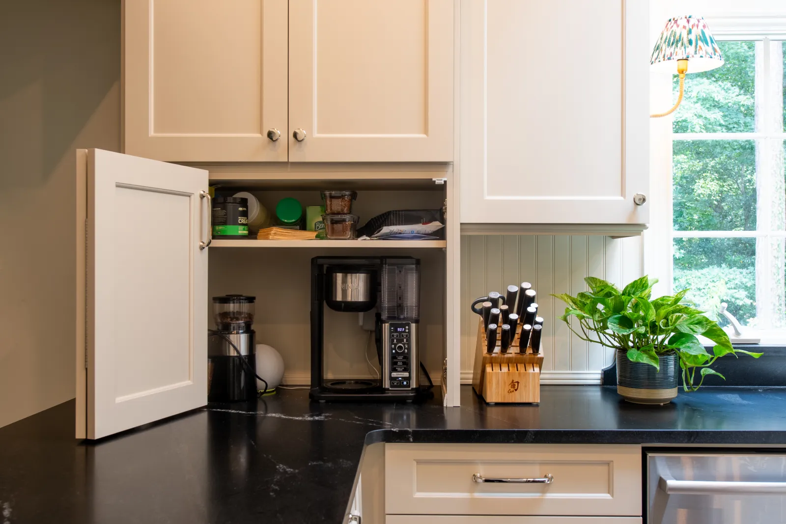 Modern kitchen countertop with open cabinet housing coffee maker, knife set, and a potted plant near a window.