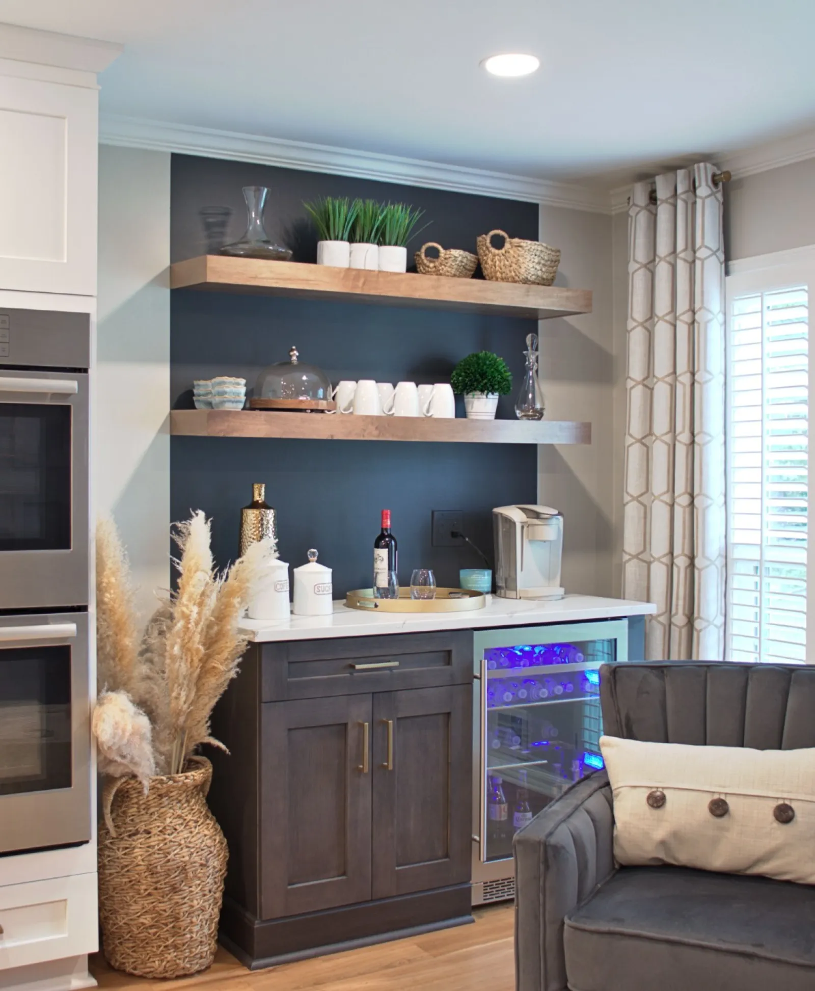 Modern kitchen corner with double stainless steel oven, dark cabinetry, open shelves, and cozy gray armchair near window shutters.
