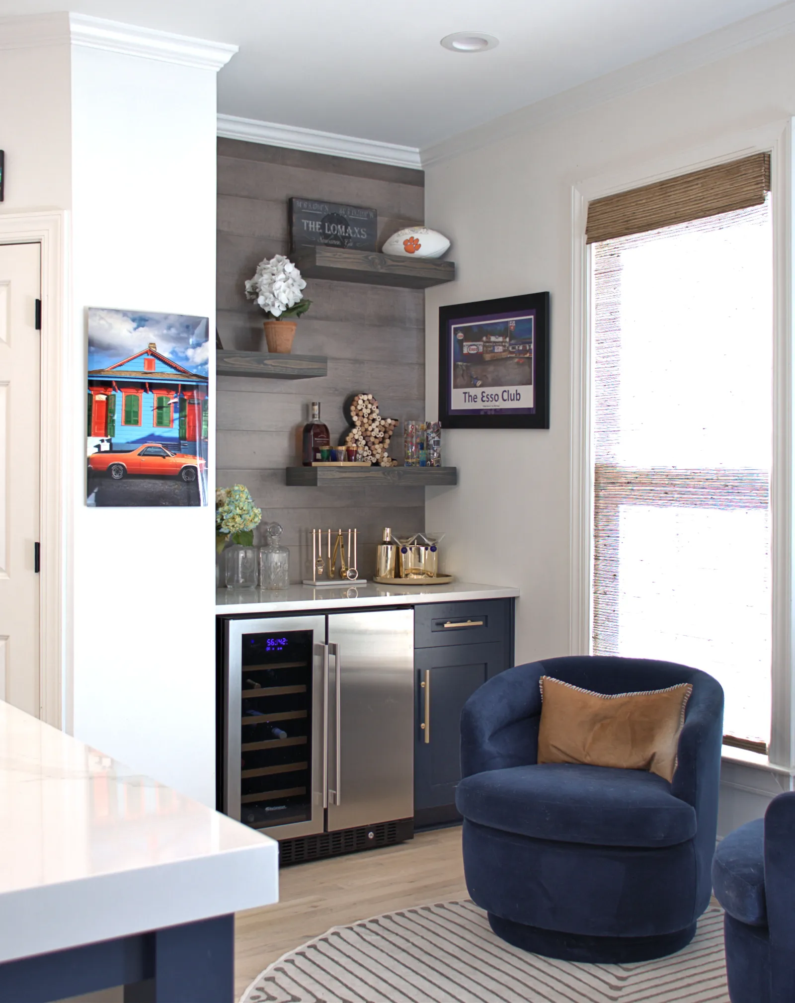 Cozy corner with blue swivel chair, built-in wine fridge, floating shelves, and framed wall art near a window with blinds.