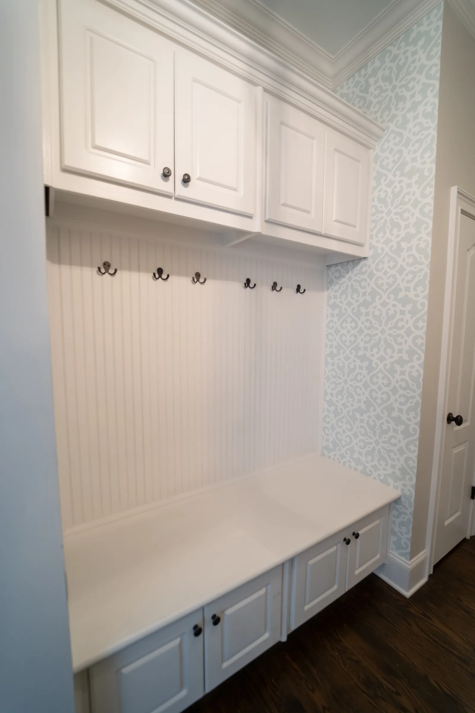 White mudroom bench with storage cabinets, wall hooks, and patterned wallpaper on wood floor.