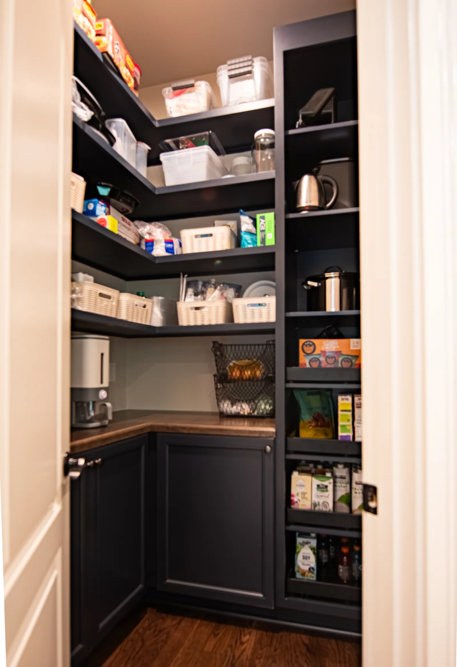 Organized pantry with dark shelves, storage baskets, kitchen appliances, and food items on wooden floor.